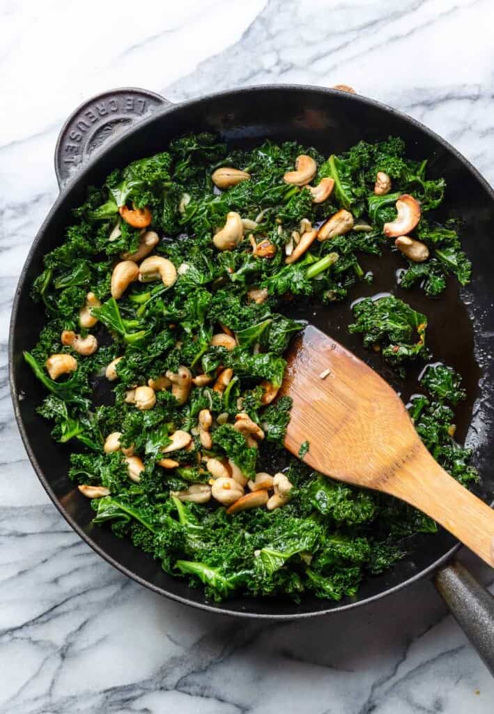Garlic & Chili Sautéed Kale with Toasted Cashews in a cast iron skillet with a wooden spoon mixing the pan