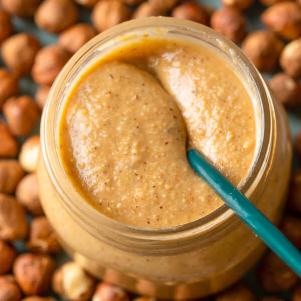 a jar of homemade hazelnut butter on a plate of hazelnuts with a spoon inside the jar