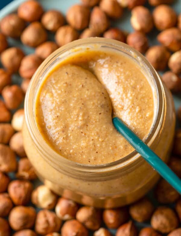 a jar of homemade hazelnut butter on a plate of hazelnuts with a spoon inside the jar