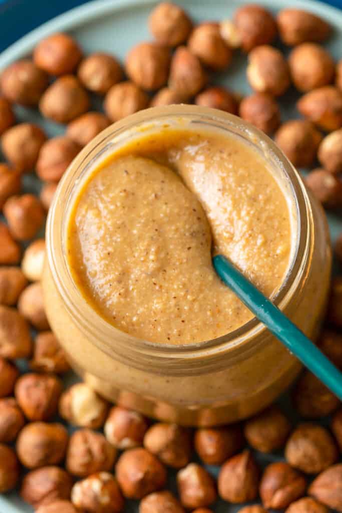 a jar of homemade hazelnut butter on a plate of hazelnuts with a spoon inside the jar