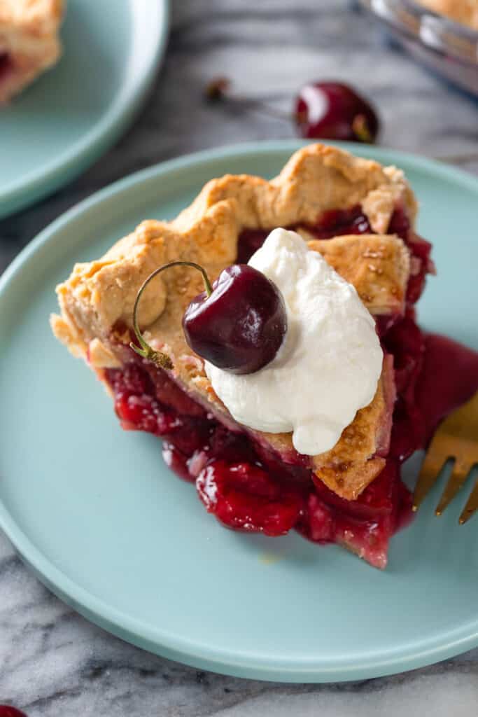 a slice of Gluten Free Cherry Pie with a dollop of whipped cream and fresh cherry on top on a cake plate