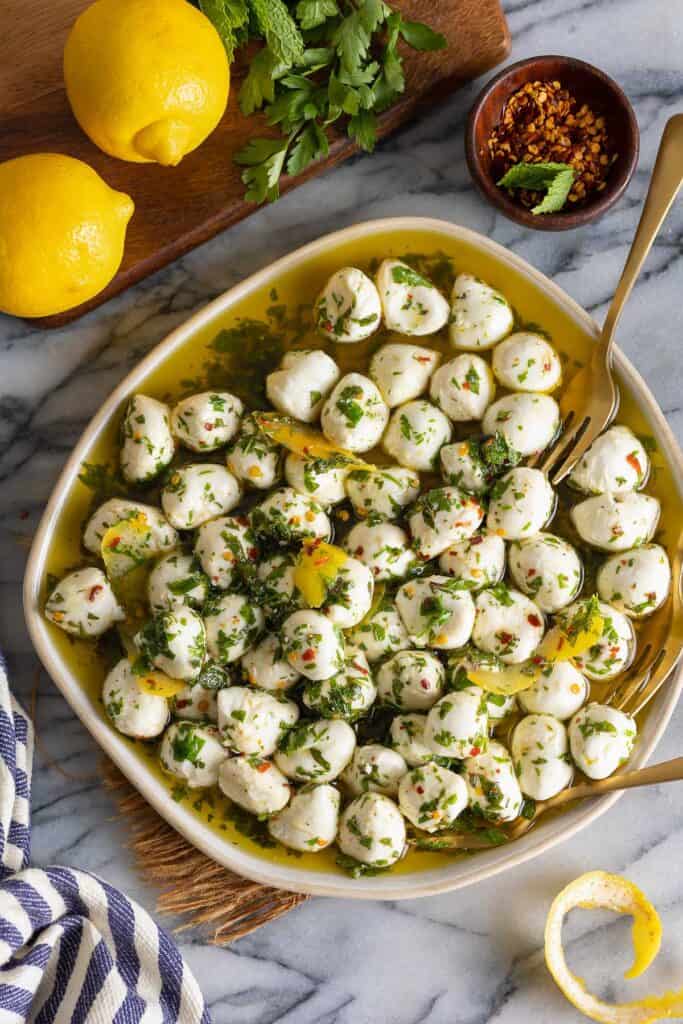 Marinated Mozzarella with Lemon Chili Herbs in a rimmed plate with mini forks along the side of the plate. A couple of lemons above the plate with a lemon peel below the plate.