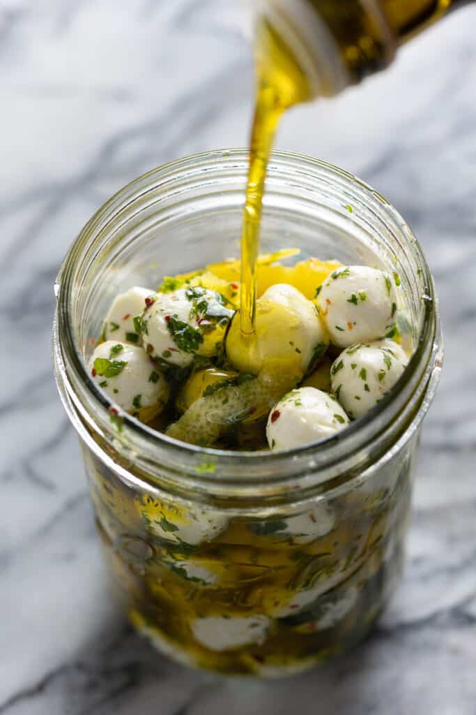 A jar full of marinated mozzarella balls with olive oil pouring over the top to cover the balls.