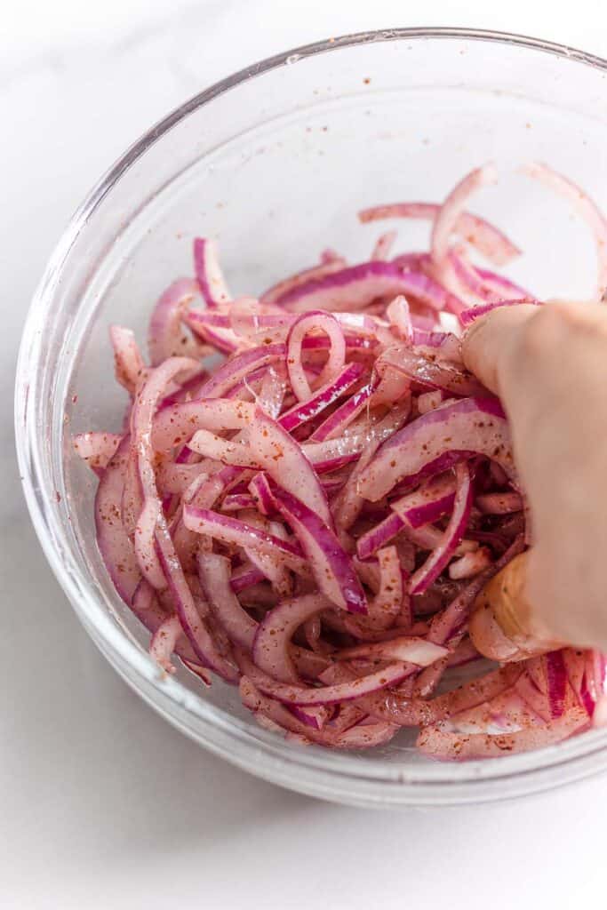 a bowl of sliced red onions, salt and sumac massaged by a hand