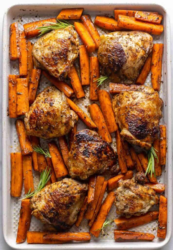 Balsamic Glazed Chicken Thighs and carrots cut into sticks on a baking sheet with rosemary sprigs