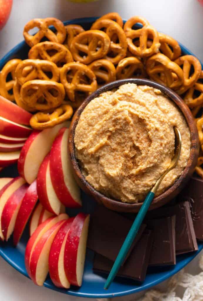 a serving plate with a bowl of apple hummus and apple slices, chocolate squares and pretzels