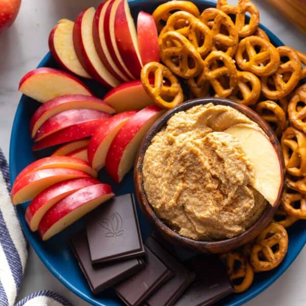 apple hummus in a bowl with an apple slice on a serving plate with more apple slices, pretzels and chocolate pieces