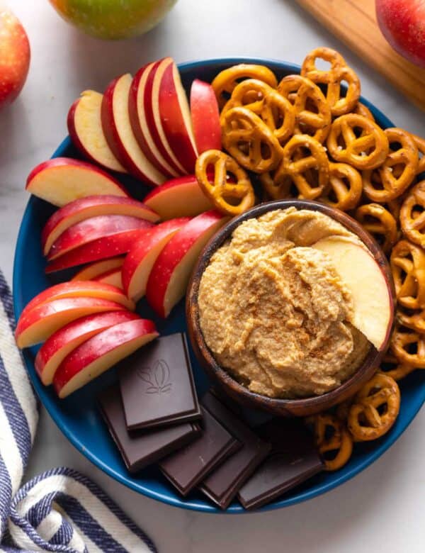 apple hummus in a bowl with an apple slice on a serving plate with more apple slices, pretzels and chocolate pieces
