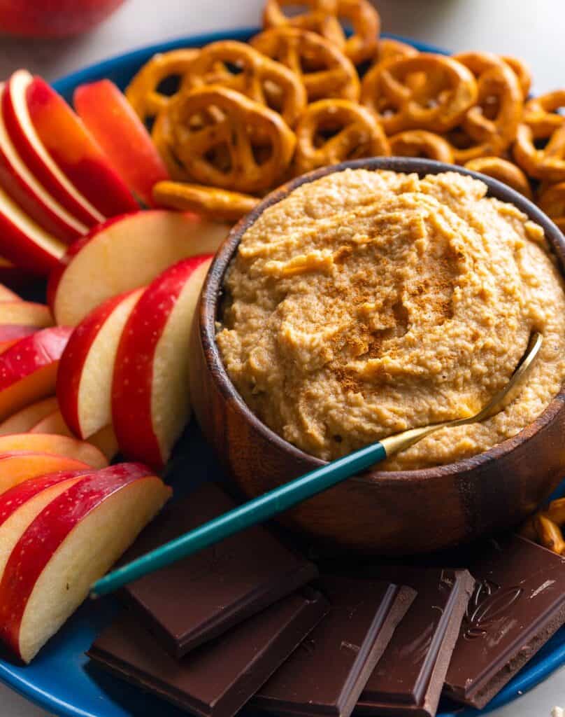 a small bowl of apple hummus with cinnamon sprinkled on top with a spoon and surrounded by apple slices, chocolate and pretzels to dip