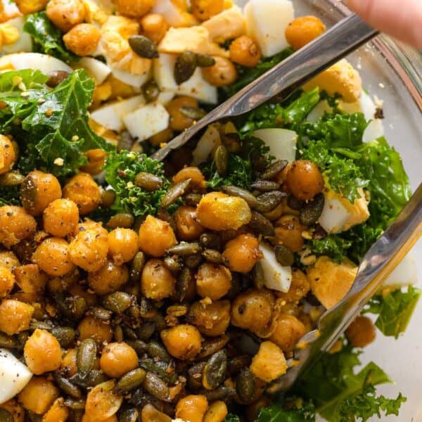 a salad made of kale, spiced chickpeas, pepitas and hard boiled egg chopped up in a bowl with a pair of tongs mixing everything together
