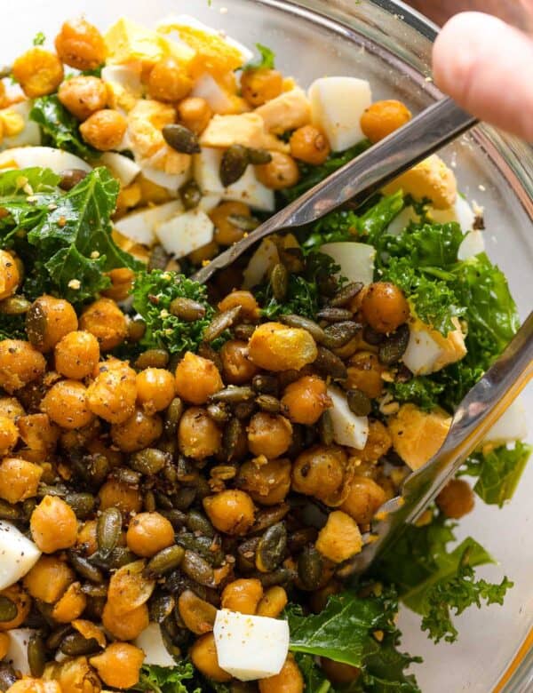 a salad made of kale, spiced chickpeas, pepitas and hard boiled egg chopped up in a bowl with a pair of tongs mixing everything together