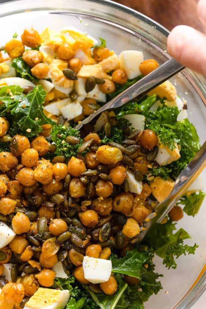 a salad made of kale, spiced chickpeas, pepitas and hard boiled egg chopped up in a bowl with a pair of tongs mixing everything together