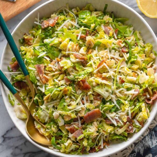 Bacon and Brussels Sprout Salad in a salad bowl with serving spoons with lemon halves and parmesan on the sides