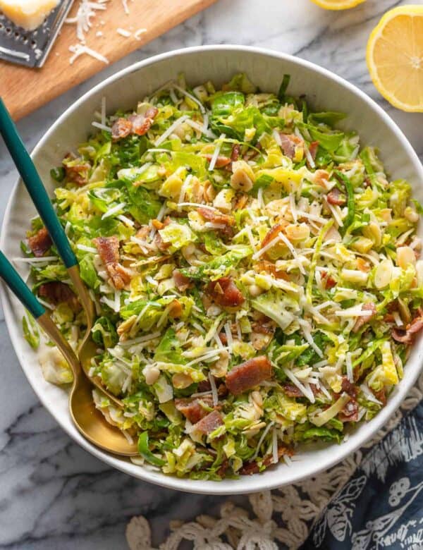 Bacon and Brussels Sprout Salad in a salad bowl with serving spoons with lemon halves and parmesan on the sides