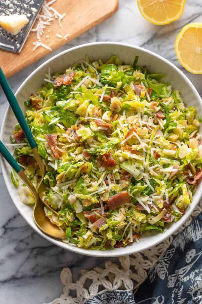 Bacon and Brussels Sprout Salad in a salad bowl with serving spoons with lemon halves and parmesan on the sides