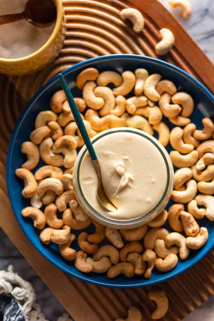 homemade cashew butter in a jar with a spoon on a plate surrounded by toasted cashews