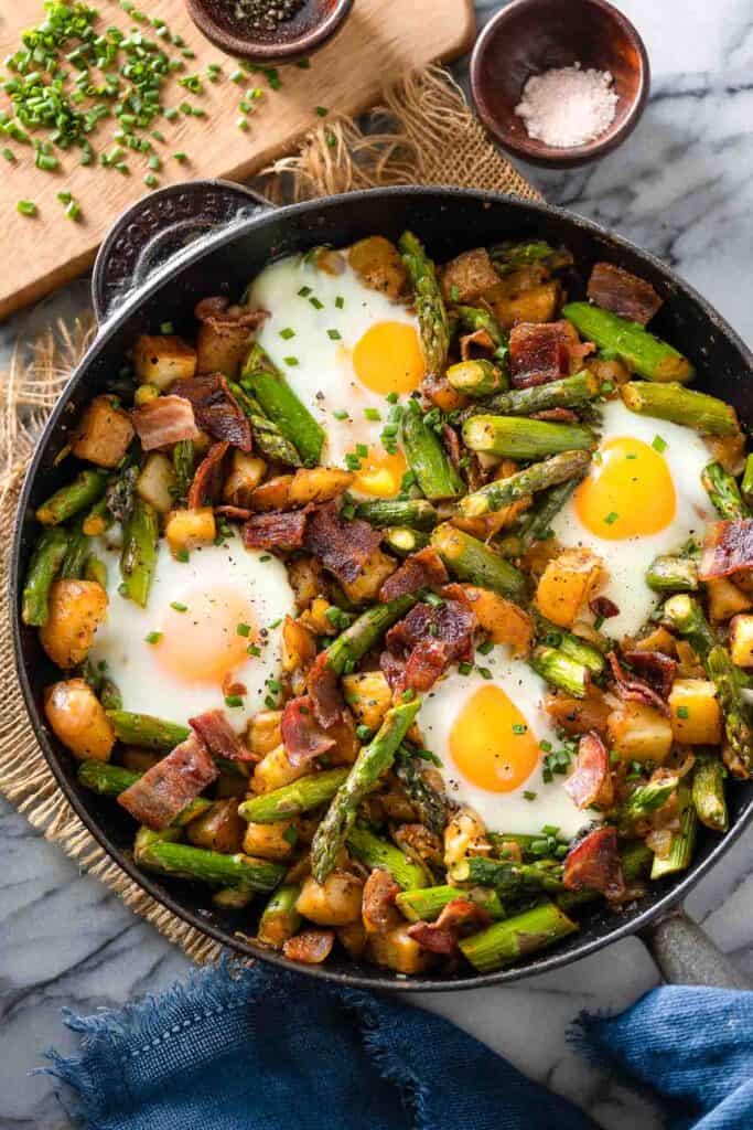 a cast iron skillet full of a Asparagus Potato Hash with Bacon & Eggs topped with fresh chopped chives