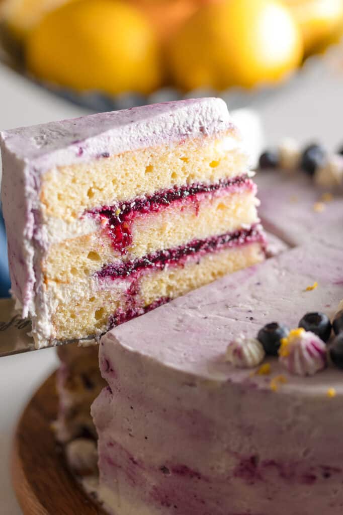 A cake knife picking up a slice of a three layer gluten free lemon cake with a blueberry compote filling and whipped mascarpone frosting