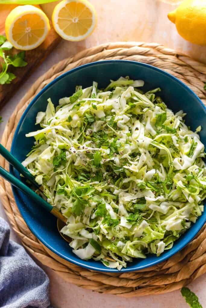 malfouf salad (lebanese cabbage slaw) in a salad bowl with a pair of serving forks surrounded by fresh lemons and herbs