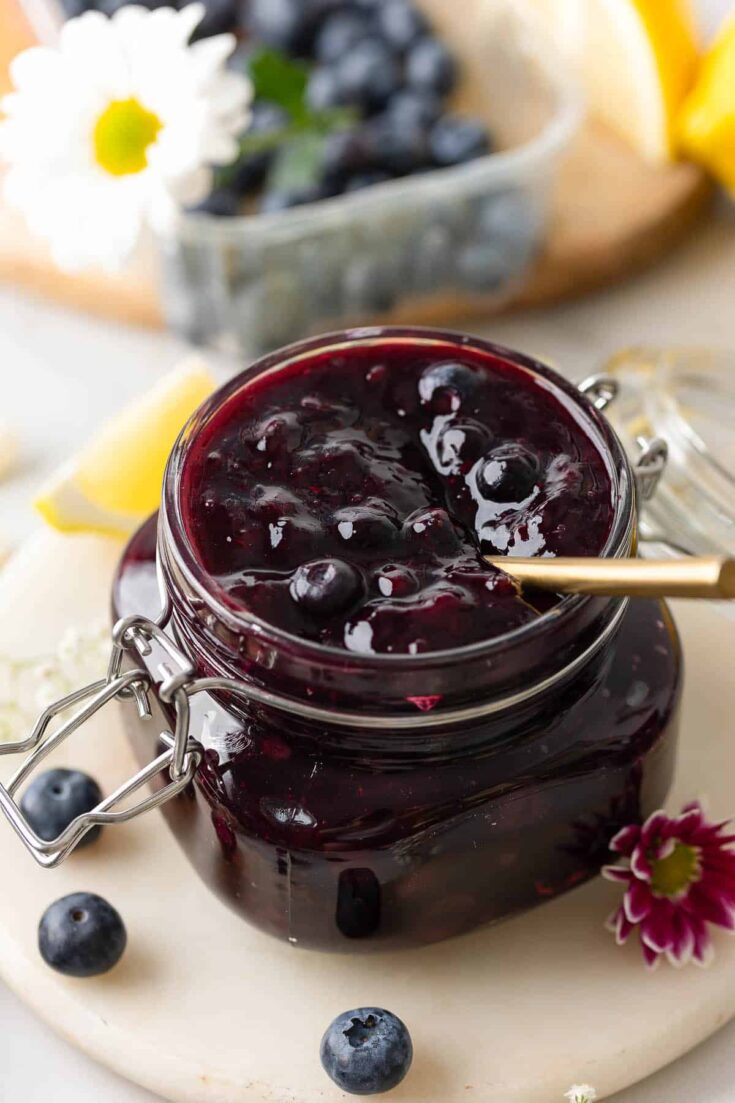homemade blueberry filling for cakes and pies in a jar with a spoon surrounded by fresh berries