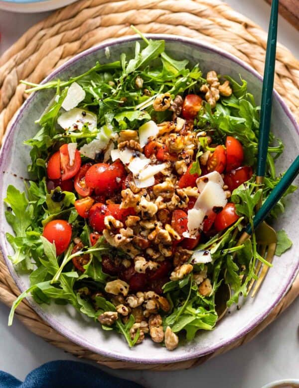 a Tomato Arugula Salad with toasted walnuts and shaved parmesan drizzled with olive oil and a balsamic vinegar reduction in a salad bowl with serving spoons