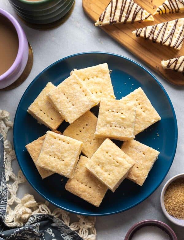 Gluten Free Shortbread cut into squares on a plate with some shortbread cookies cut into triangles and drizzled with chocolate and a couple mugs on coffee on the side