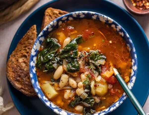 A bowl full of Bacon Kale Bean Soup spoon on a plate with a couple slices of gluten free toast; chili flakes and black pepper in small bowls on the side