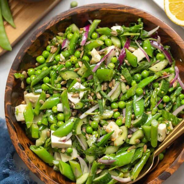 A Spring Greens Salad in a bowl made with snap peas, english peas, asparagus, cucumber, onion, mozzarella and sunflower seeds with the fresh ingredients surrounding the bowl.