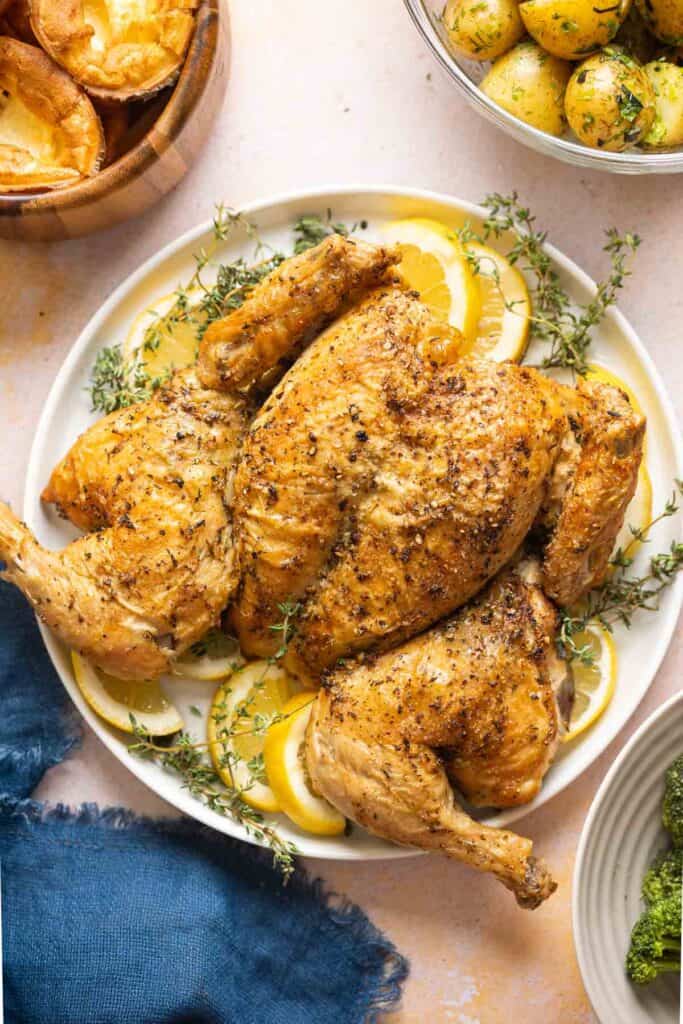 Spatchcocked Chicken on a plate with fresh thyme and lemon slices with herbed potatoes and yorkshire puddings on the side.