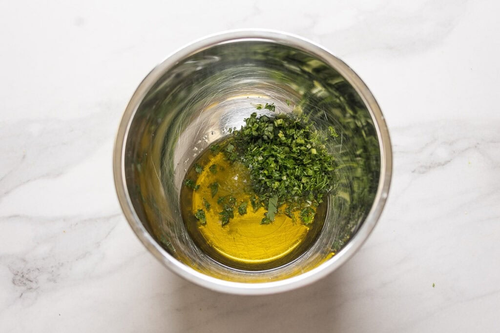 A mixing bowl with olive oil and chopped fresh herbs inside.