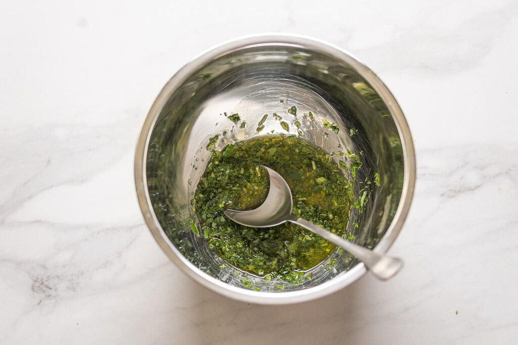 A mixing bowl with olive oil and chopped fresh herbs mixed together with a spoon.