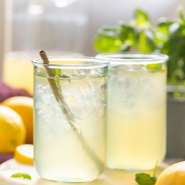 Two glasses full of iced Basil Lemonade with fresh basil leaves and lemons on the side.