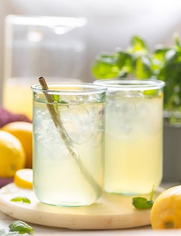 Two glasses full of iced Basil Lemonade with fresh basil leaves and lemons on the side.