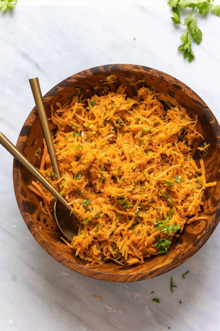 A French Carrot Salad in a salad bowl with a serving spoon and fork with fresh parsley on the side.