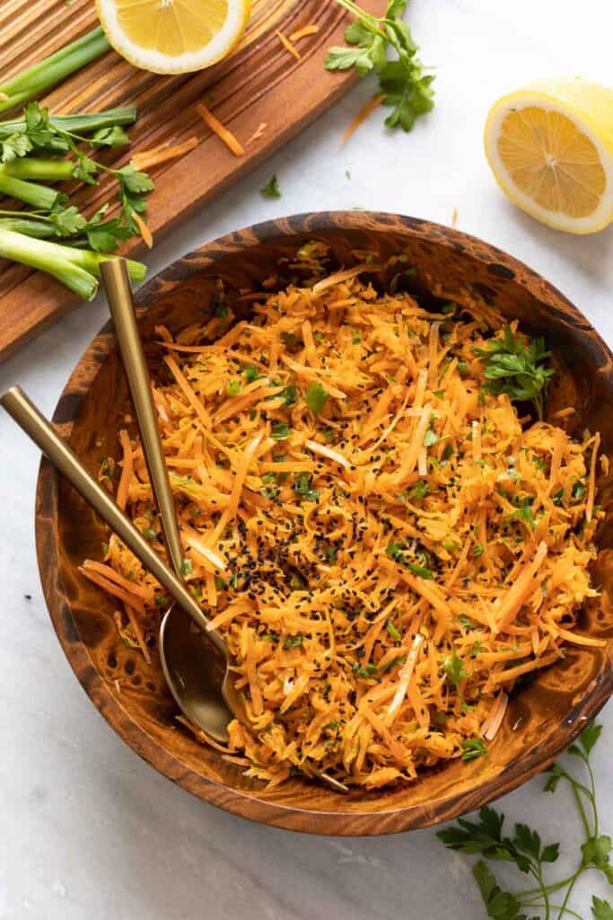 A French Carrot Salad garnished with black sesame seeds in a salad bowl with a serving spoon and fork and the fresh ingredients around the bowl.