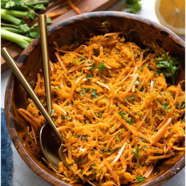A French Carrot Salad in a salad bowl with a serving spoon and fork and the fresh ingredients around the bowl (spring onion, lemon, parsley).