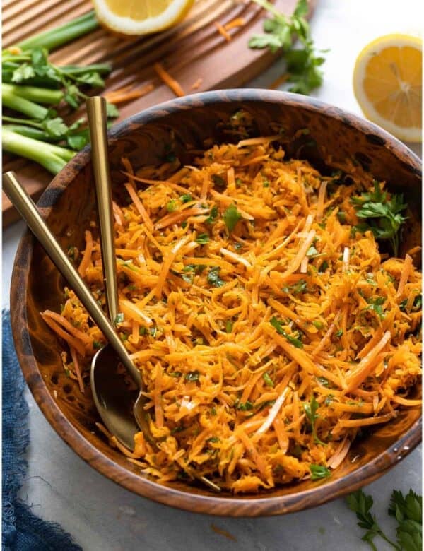 A French Carrot Salad in a salad bowl with a serving spoon and fork and the fresh ingredients around the bowl (spring onion, lemon, parsley).
