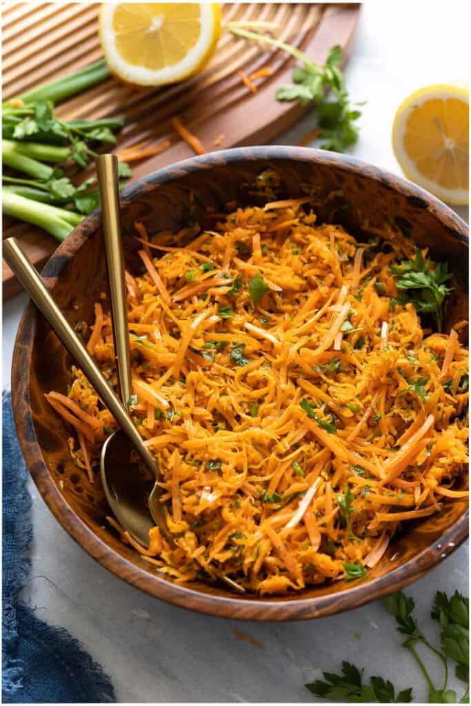 A French Carrot Salad in a salad bowl with a serving spoon and fork and the fresh ingredients around the bowl (spring onion, lemon, parsley).
