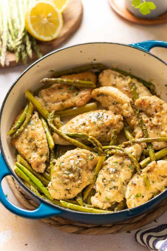 Gluten free breaded and pan fried lemon chicken with asparagus in a large pan; fresh asparagus, lemon and parsley on the side.