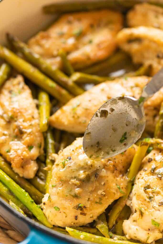 A spoon dripping lemon sauce back onto a breaded and pan fried chicken breast in a pan with asparagus.