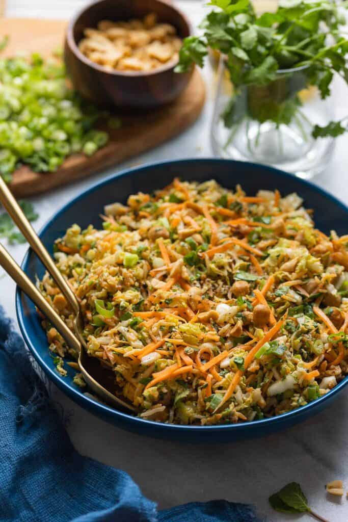 A salad bowl with serving utensils full of a cabbage crunch salad: shredded cabbage, carrots, spring onion, herbs and peanuts in a peanut dressing. Fresh cilantro, chopped peanuts and spring onions in the back.