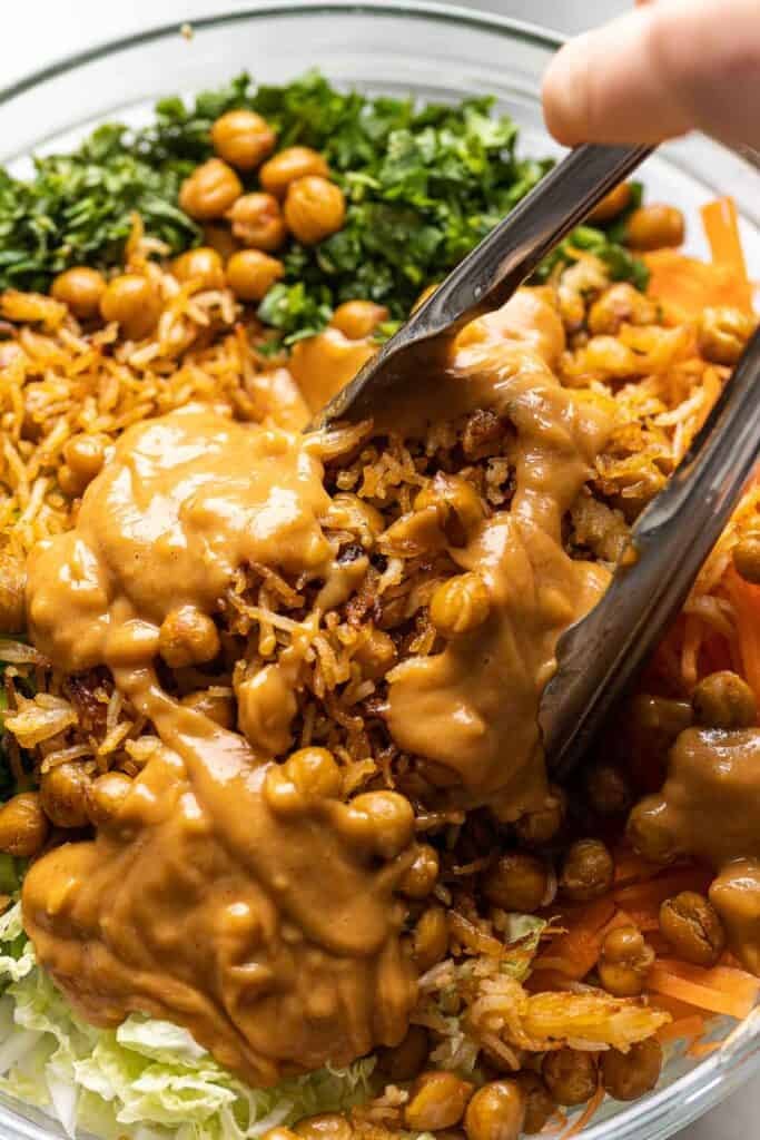 A pair of kitchen tongs mixing up a cabbage crunch salad with a creamy peanut butter dressing.