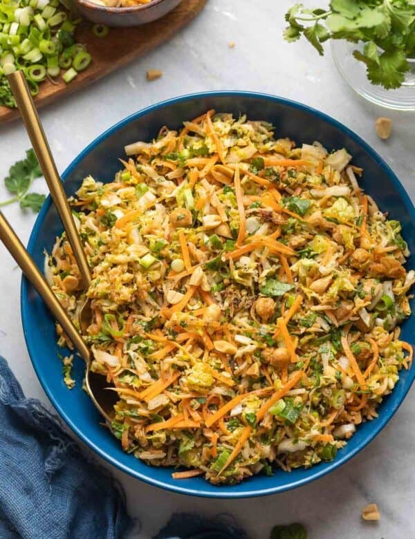 A salad bowl with serving utensils full of a Crispy Peanut Rice & Cabbage Crunch Salad: shredded cabbage, carrots, spring onion, herbs and peanuts in a peanut dressing.