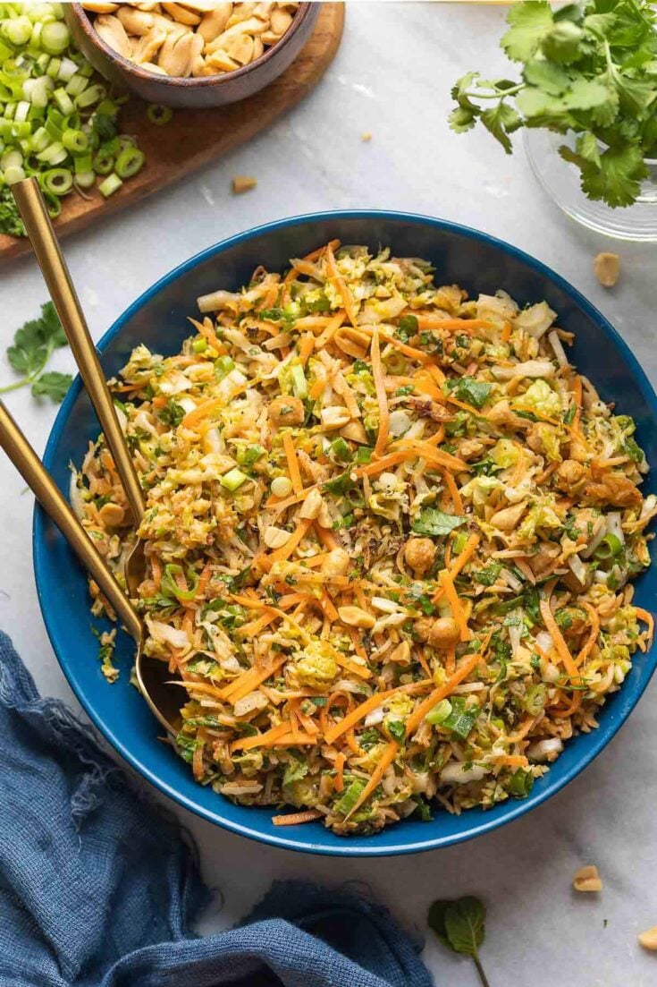 A salad bowl with serving utensils full of a Crispy Peanut Rice & Cabbage Crunch Salad: shredded cabbage, carrots, spring onion, herbs and peanuts in a peanut dressing.