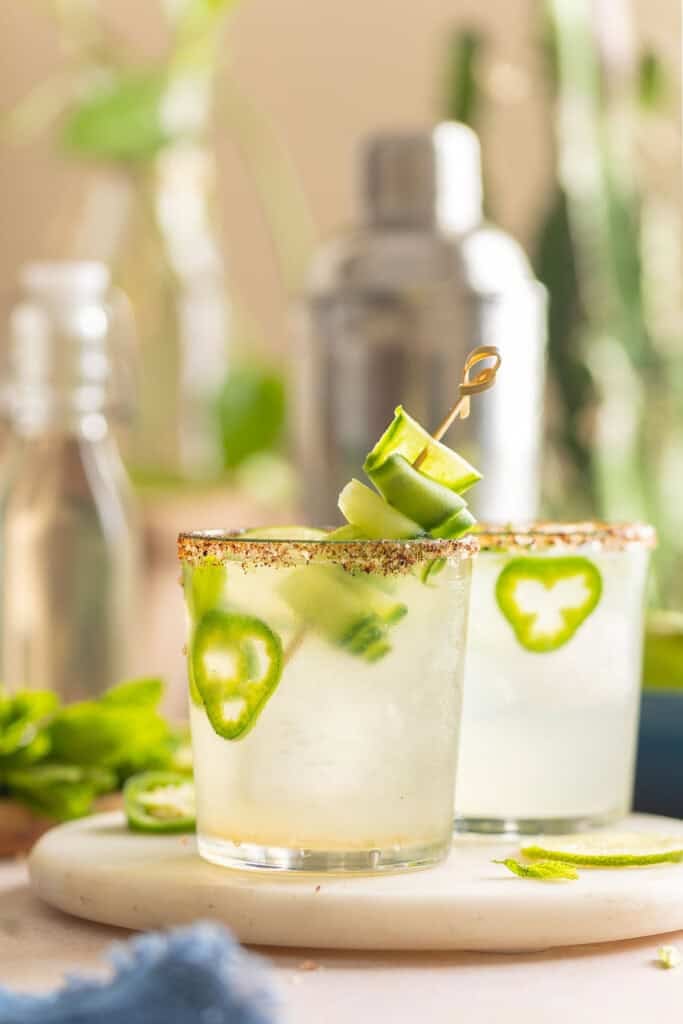 Two glasses of Jalapeño Cucumber Margarita in cups rimmed with a salt and chili powder mixture and cucumber slices, jalapeno slices and fresh mint leaves inside
