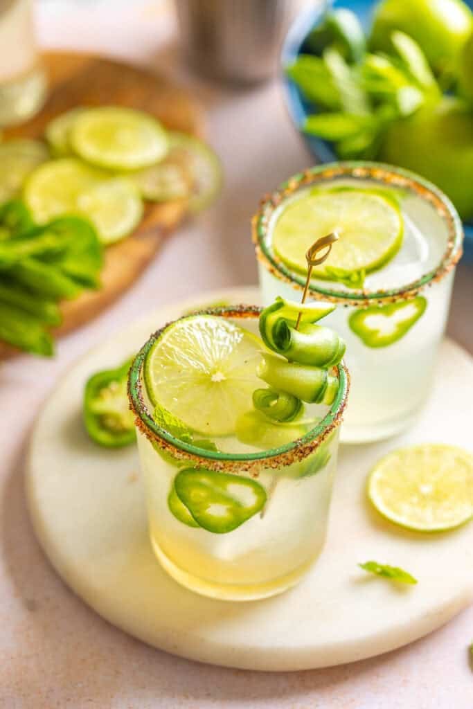 A couple glassed of Jalapeño Cucumber Margarita garnished with jalapeno slices, cucumber slices, fresh mint leaves and salt/chili rimmed glasses.