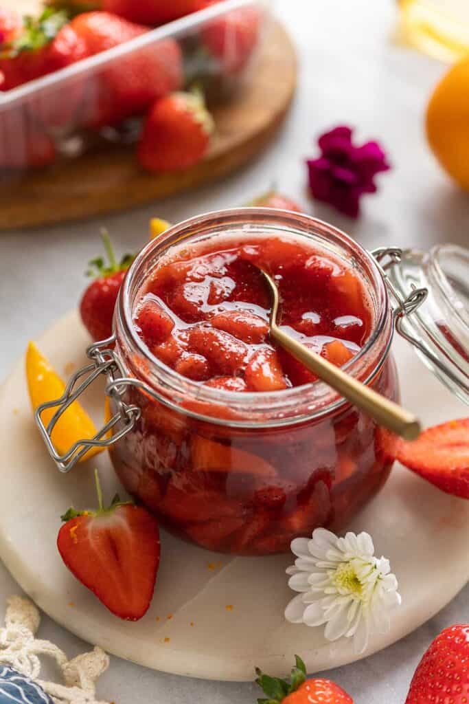 A jar of homemade strawberry compote surrounded by fresh strawberries orange slices and flowers.
