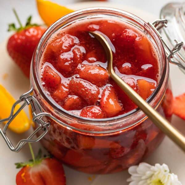 A jar of homemade strawberry compote with a spoon in the jar surrounded by fresh strawberries & orange slices.