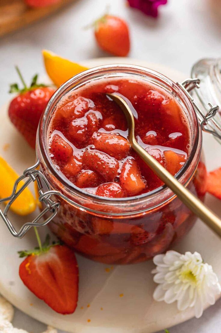 A jar of homemade strawberry compote with a spoon in the jar surrounded by fresh strawberries & orange slices.