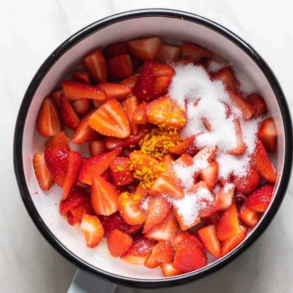 A saucepan full of quartered strawberries, sugar, orange juice and zest.